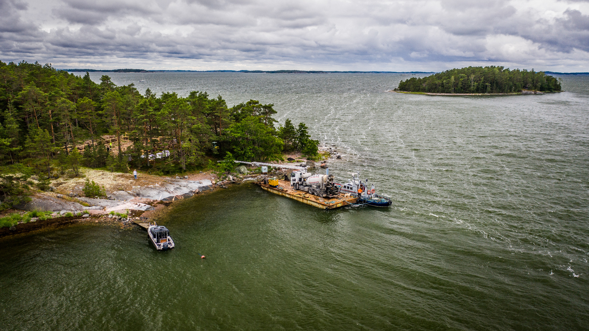 Saaren ranta, jossa proomukuljetus betoniautolla, useita aluksia, itämeri, metsää ja pilvinen taivas.